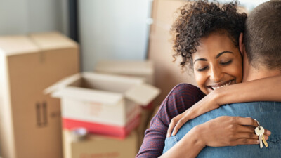 New homeowners embracing, holding house keys, in front of a pile of moving boxes.