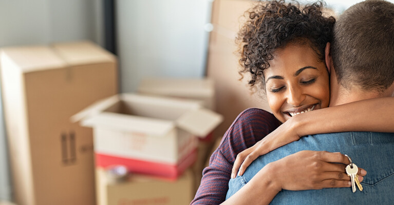 New homeowners embracing, holding house keys, in front of a pile of moving boxes.