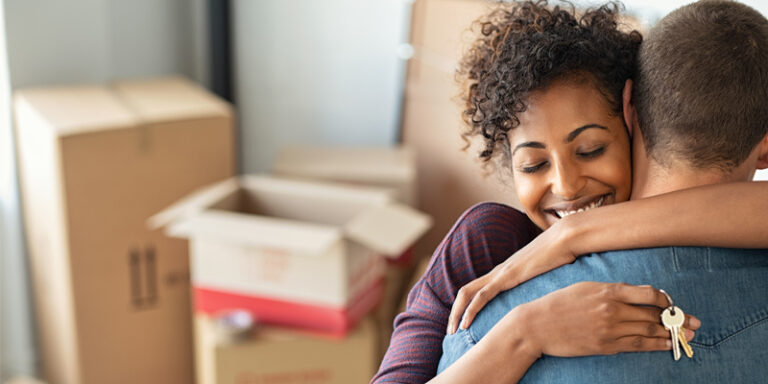 New homeowners embracing, holding house keys, in front of a pile of moving boxes.