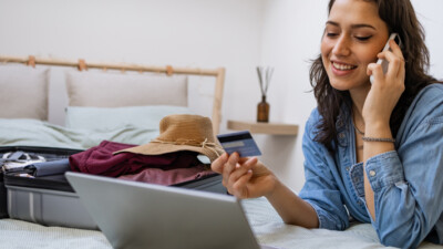 A woman speaking on her cell phone, holding her credit card, and looking at a laptop. There is a suitcase of vacation clothing on the bed.