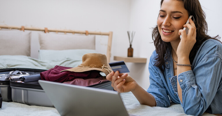 A woman speaking on her cell phone, holding her credit card, and looking at a laptop. There is a suitcase of vacation clothing on the bed.