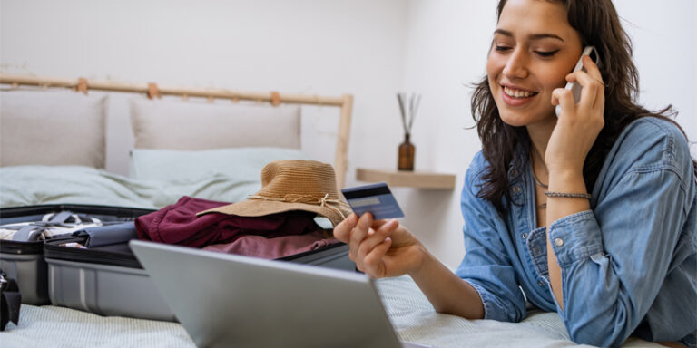 A woman speaking on her cell phone, holding her credit card, and looking at a laptop. There is a suitcase of vacation clothing on the bed.
