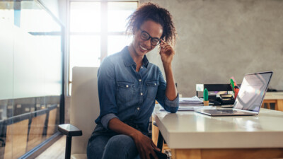 A woman sitting at her desk in an office, smiling and adjusting her glasses.