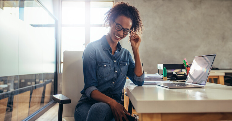 A woman sitting at her desk in an office, smiling and adjusting her glasses.