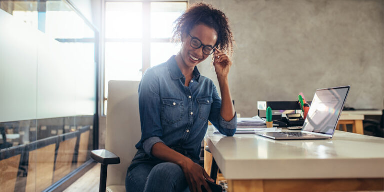 A woman sitting at her desk in an office, smiling and adjusting her glasses.
