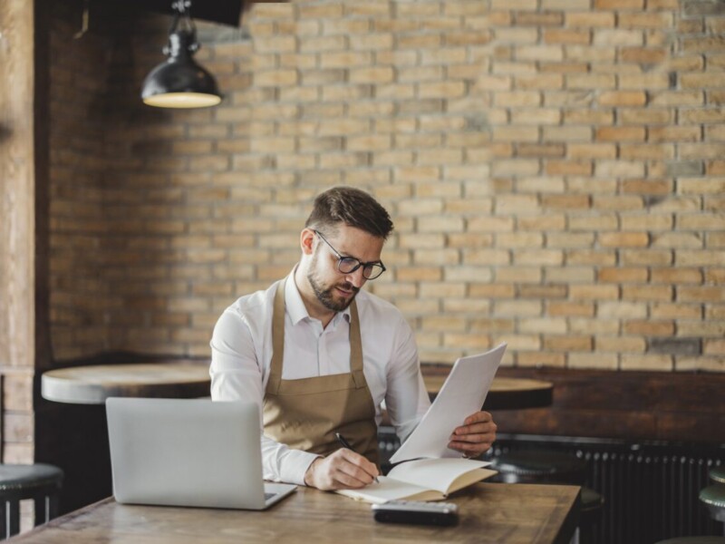 Man wearing apron at a cafe, using a laptop and looking at documents.