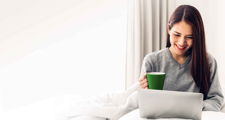 A woman sitting in bed with her laptop, holding a green mug.