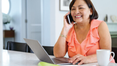 A woman smiling and using her laptop at her kitchen table, while speaking on the phone.