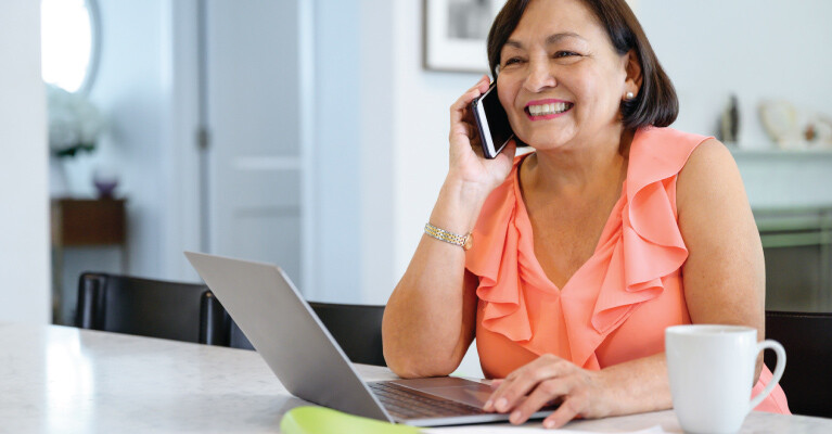 A woman smiling and using her laptop at her kitchen table, while speaking on the phone.