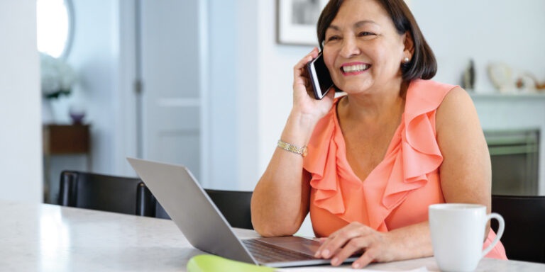 A woman smiling and using her laptop at her kitchen table, while speaking on the phone.