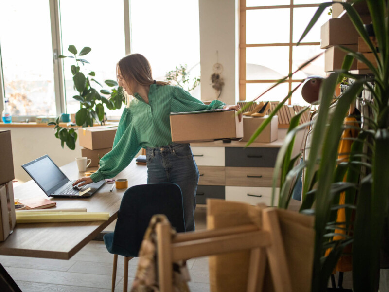 Businesswoman holding box, doing shop inventory on a laptop.