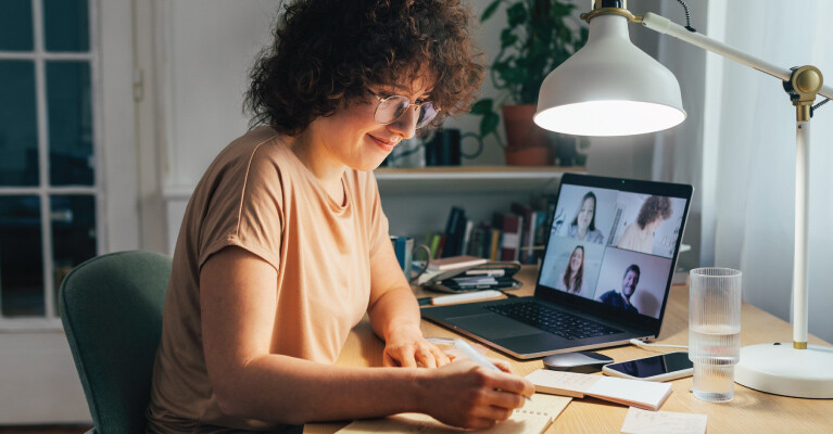 A woman writing in a notebook while on a video call with other people.