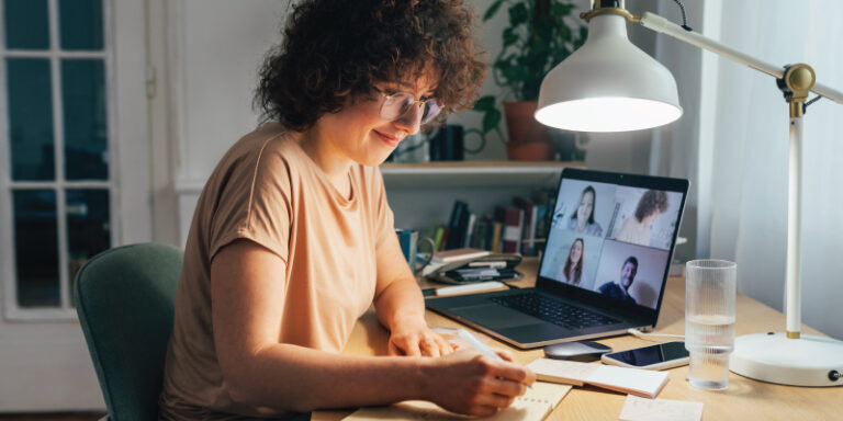 A woman writing in a notebook while on a video call with other people.