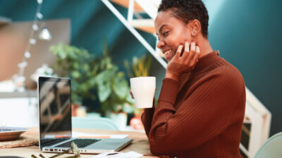 A woman drinking from a mug and smiling at her laptop.