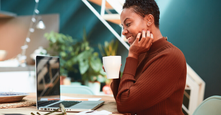 A woman drinking from a mug and smiling at her laptop.