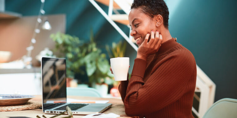 A woman drinking from a mug and smiling at her laptop.