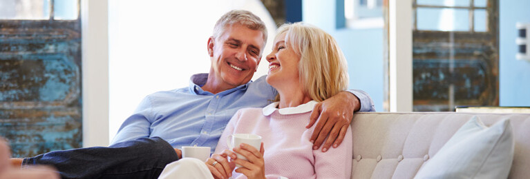 A man and a woman sitting on a couch, laughing and holding mugs.