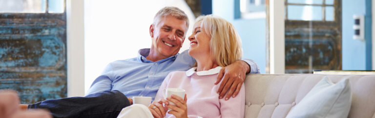 A man and a woman sitting on a couch, laughing and holding mugs.