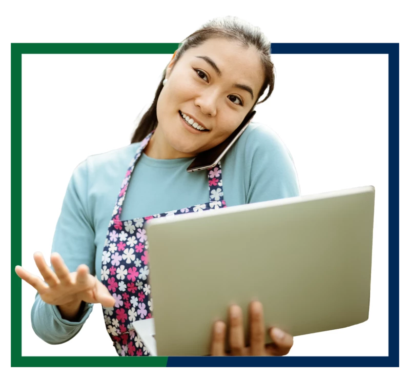 A business owner wearing a floral apron and holding a laptop, while talking on her cell phone.