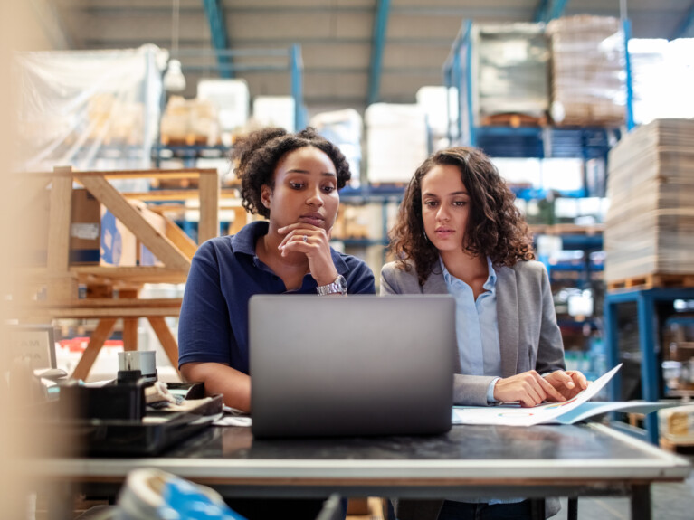 Two women in a warehouse using a laptop.
