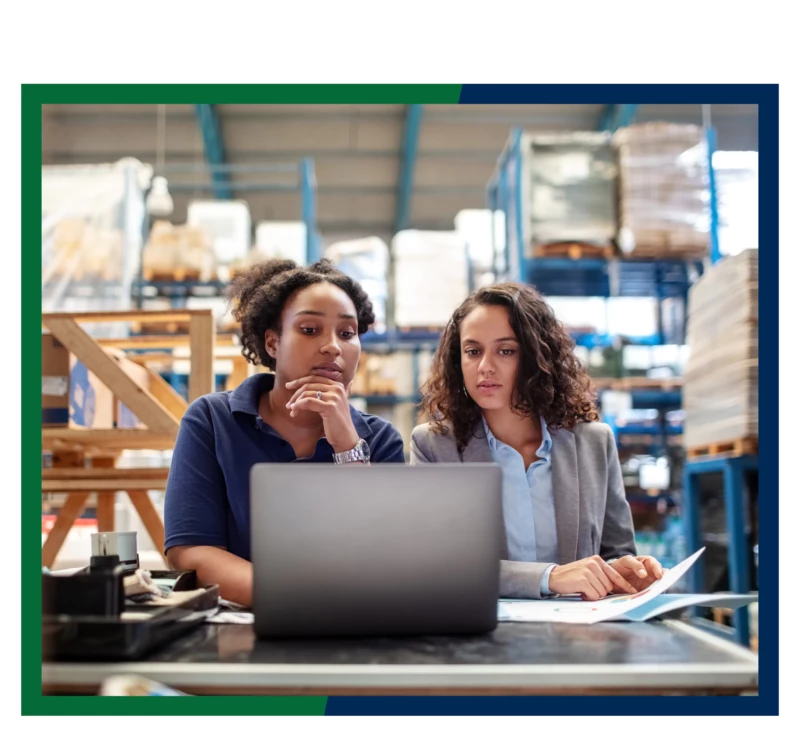 Two women in a warehouse using a laptop.